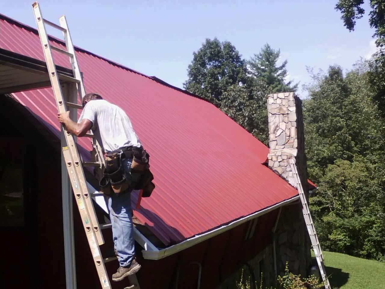 Roofer on a ladder beside a red metal roof and stone chimney — Soffit & Fascia Repair by Roofing Brothers in Smithville